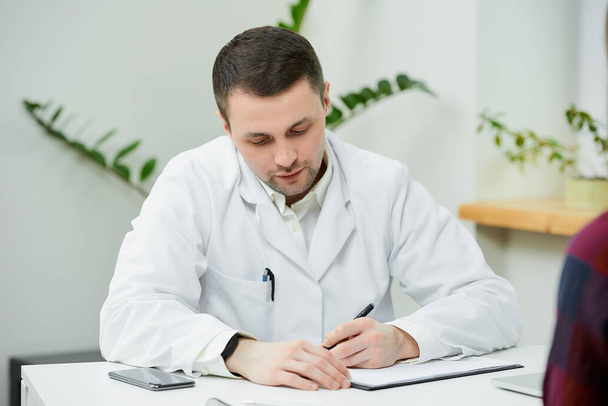 A portrait of a doctor in a white lab coat who is sitting at a desk and writing a diagnosis to a female patient in a doctor's office. A woman at a doctor's appointment in a hospital. - Foto, Imagen