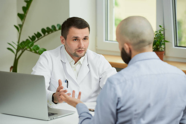 A caucasian doctor in a white lab coat is sitting at a desk and describing the treatment of a bald male patient in a hospital. A man with a beard at an appointment in a doctor's office. - Fotoğraf, Görsel