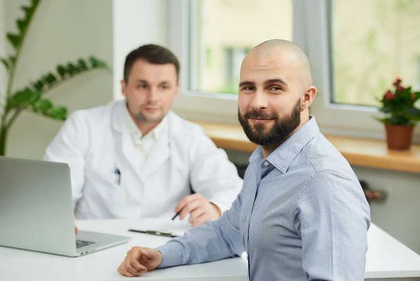 A bald male patient is rejoicing in the success of his healing near a caucasian doctor in a white lab coat in a hospital. A happy man with a beard at an appointment in a doctor's office. - Photo, Image