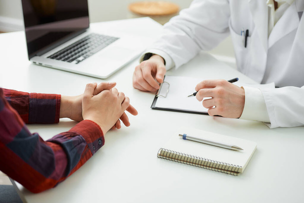 Egy fotó egy olyan beteg összecsukott kezéről, aki egy orvos közelében gesztikulál egy kórházban. Egy jegyzetfüzet tollal, írótáblával, laptoppal egy fehér asztalon fekszik egy orvosi rendelőben. - Fotó, kép