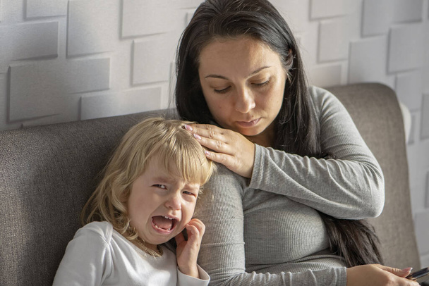 Una madre amorevole si accarezza la testa e calma la sua piccola figlia che piange mentre è seduta sul divano - Foto, immagini