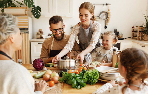 Positive young housewife with little daughters, husband and grandmother gathering around kitchen table and preparing delicious healthy dishes with fresh ingredients for family lunch at hom - Photo, Image