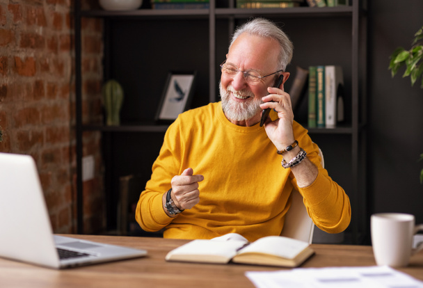 Tyylikäs iäkäs mies hymyilee ja vastaa puheluun istuessaan työpöydän ääressä kodikkaassa työpaikassa hom - Valokuva, kuva