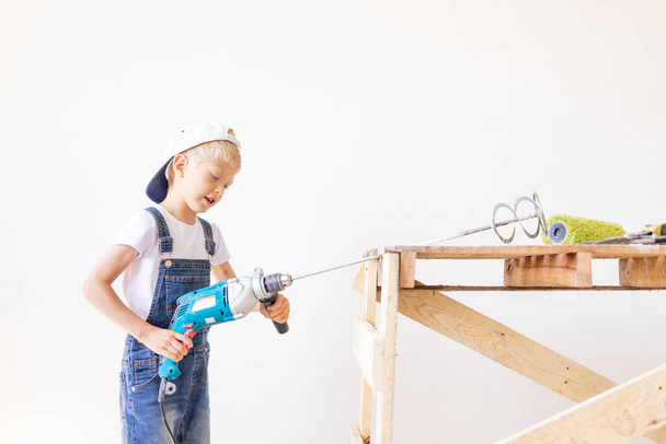 un bambino costruttore tiene un miscelatore di costruzione sullo sfondo di una parete bianca e una scala di costruzione, un luogo per il testo, un concetto di costruzione e riparazione - Foto, immagini