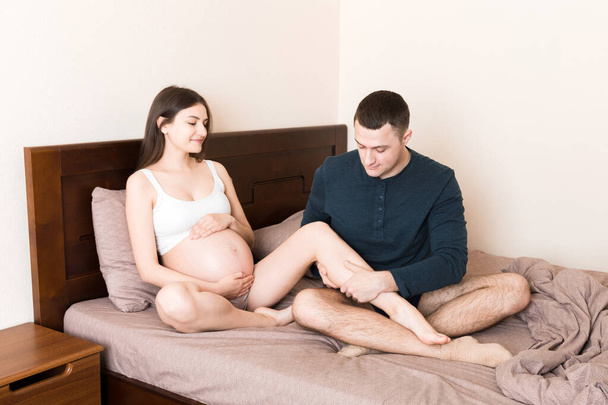 Счастливая беременная семья. Муж массирует ноги беременной жене и сидит вместе на диване в их доме. - Фото, изображение