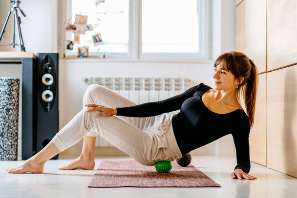 Urocza brunetka używająca piłeczki do masażu z punktu zapłonu do samodzielnego masażu z zastosowaniem techniki łagodzenia bólu pleców i miednicy, ćwicząca na macie fitness w domu na tle. - Zdjęcie, obraz