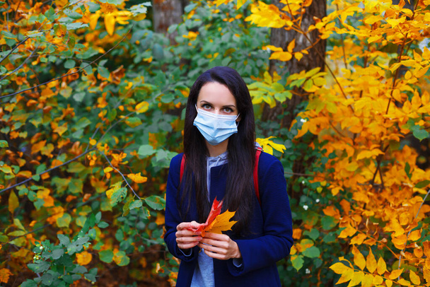 Όμορφη γυναίκα σε προστατευτική μάσκα κρατώντας φύλλα σφενδάμου σε πολύχρωμο δάσος του φθινοπώρου. Νέα κανονική έννοια. Χρόνος παραμονής στο ύπαιθρο κατά τη διάρκεια της πανδημίας covid 19 - Φωτογραφία, εικόνα