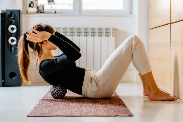 Zijaanzicht vrouw doet zelf-massage techniek toepassen van speciale duoball of bal voor rugpijn verlichting, uit te werken op fitness mat op de vloer met mat thuis interieur op de achtergrond. - Foto, afbeelding