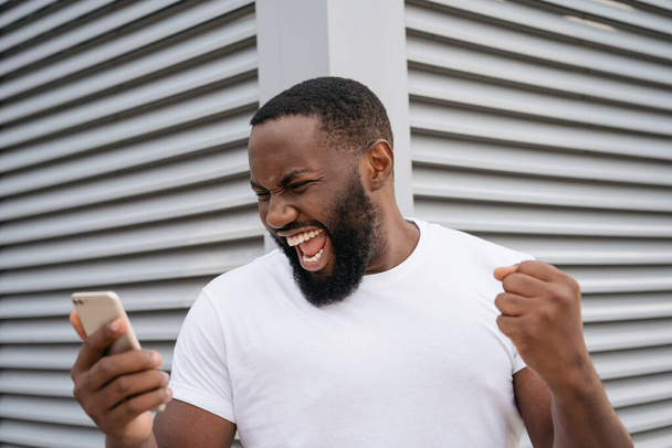 Portrait of overjoyed online lottery winner looking at digital screen, celebration success. Sport betting concept. Excited African American man using mobile phone, shopping online on Black Friday - Photo, Image