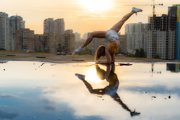 Flexible female gymnast doing handstand and calisthenic with reflection in the water on cityscape background during dramatic sunset - Foto, afbeelding