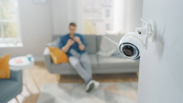 Закройте снимок современной камеры наблюдения Wi-Fi с двумя антеннами на белой стене в уютном помещении. Человек сидит на диване на заднем плане. - Фото, изображение