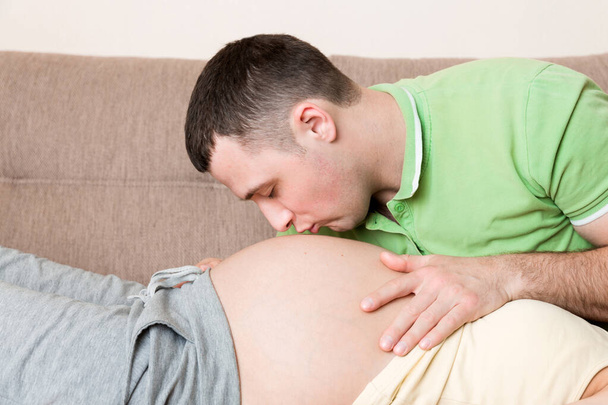 Молодой бородатый мужчина прижимает ухо к животу беременной жены и целует его.. - Фото, изображение