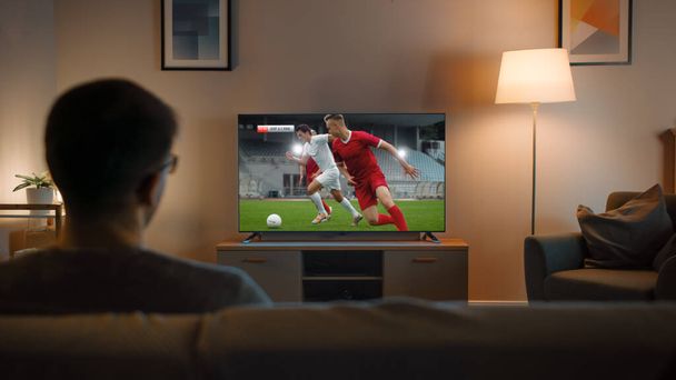 Young Man in Glasses is Sitting on a Sofa and Watching TV with a Soccer Match. Its Evening and Room at Home Has Working Lamps. - Фото, изображение