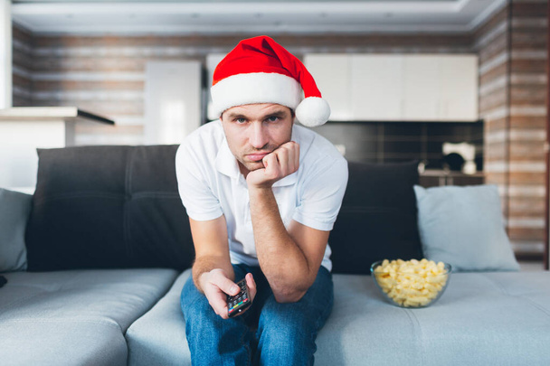 Ο νεαρός βλέπει τηλεόραση στο διαμέρισμά του. Βαριέται ο τύπος με το καπέλο των Χριστουγέννων να χρησιμοποιεί τηλεχειριστήριο. Δυστυχισμένο μπερδεμένο άτομο που γιορτάζει την Πρωτοχρονιά ή τα Χριστούγεννα. Κάτσε μόνος στον καναπέ.. - Φωτογραφία, εικόνα