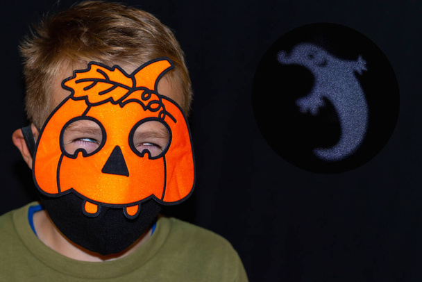 Vista frontal fechar decorações de Halloween na nova realidade da pandemia COVID-19. menino em abóbora laranja e máscara médica.silhueta branca de fantasma engraçado no fundo escuro - Foto, Imagem