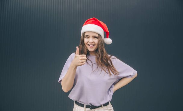 Young stylish trendy woman isolated over grey blue background. Cheerful positive girl in red festive hat hold big thumb up. Like sign or symbol. Winter time for christmas or new year period. - 写真・画像