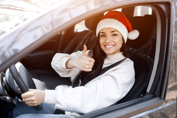 Mutlu yıllar ve mutlu Noeller. Bir kadın arabada oturur, kırmızı bir Santaclaus şapkası takar ve başparmağını kaldırır. - Fotoğraf, Görsel