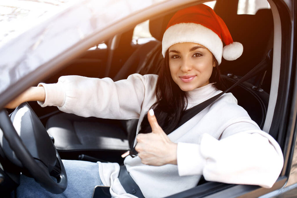 Hyvää uutta vuotta ja hyvää joulua Nainen istuu autossa, hän on pukeutunut punaiseen joulupukin hattuun ja näyttää peukalon ylös. - Valokuva, kuva