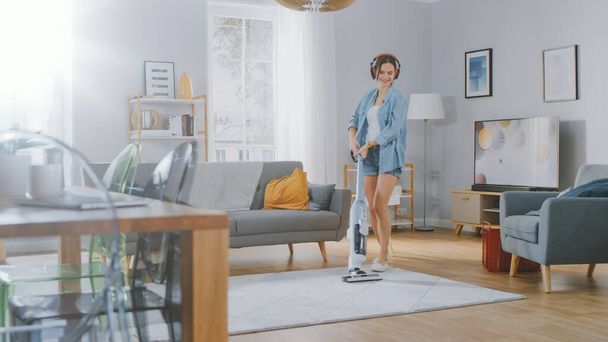 Young Beautiful Woman in Jeans Shirt and Shorts is Listening to Music on Her Headpones, Dancing and Vacuum Cleaning a Carpet in a Cozy Room at Home. She Uses a Cordless Vacuum. Shes Happy and Joyful. - Fotografie, Obrázek