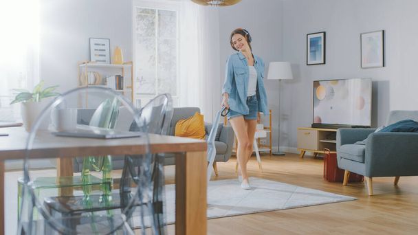 Young Beautiful Woman in Jeans Shirt and Shorts is Listening to Music on Her Headpones, Dancing and Vacuum Cleaning a Carpet in a Cozy Room at Home. She Uses a Cordless Vacuum. Shes Happy and Joyful. - Zdjęcie, obraz