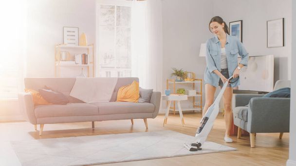 Young Beautiful Woman in Jeans Shirt and Shorts is Vacuum Cleaning a Carpet in a Bright Cozy Room at Home. She Uses a Modern Cordless Vacuum. Shes Happy and Cheerful. Shot with Warm Sun Flare. - Foto, Imagen