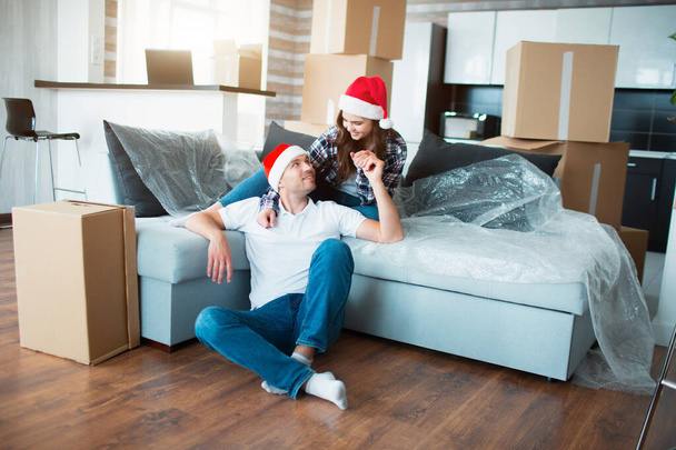 Newlywed couple celebrate Christmas or New Year in their new apartment. Young happy man and woman celebrating moving to new home and sitting among boxes. - Photo, Image