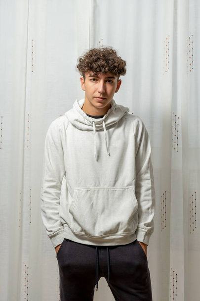 Attractive young man with curly hair wearing white sweatshirt posing on white curtains background. Fashion concept - Photo, Image