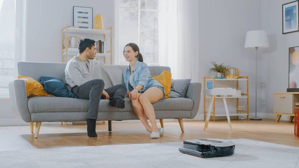 Smart Robot Vacuum Cleaner Sucking Up Dust from a Carpet. Beautiful Couple is Sitting on a Sofa and Talking in the Background. Technological Home Appliance Device Moves Past Them. - Foto, afbeelding