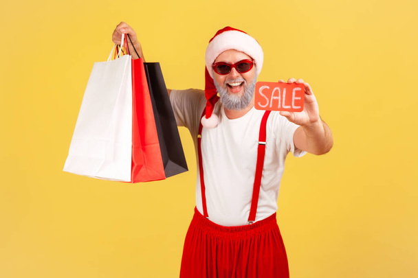Satisfied grey bearded man in stylish sunglasses and santa claus costume holding and showing paper shopping bags and sale card, holidays discounts. Indoor studio shot isolated on yellow background - Фото, изображение