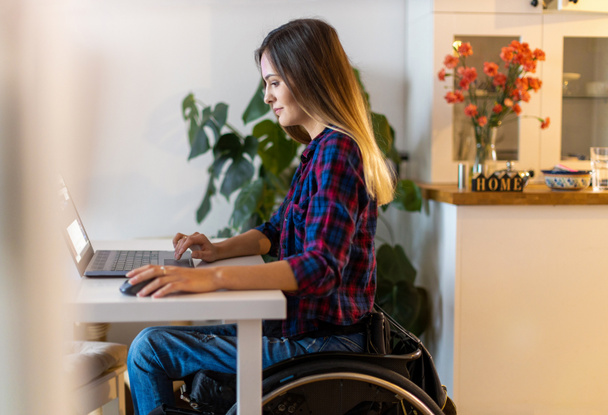 Молодая женщина в инвалидной коляске с помощью ноутбука дома - Фото, изображение