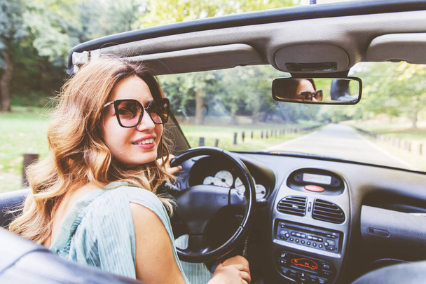 Όμορφη νεαρή γυναίκα που οδηγεί κάμπριο. Ευτυχισμένο κορίτσι οδηγεί την καλοκαιρινή μέρα. Φορώντας γυαλιά ηλίου. Πορτρέτο της χαμογελαστής γυναίκας στο αυτοκίνητο. Κοιτάζοντας την κάμερα.Αντανάκλαση στον καθρέφτη οδήγησης του αυτοκινήτου. Γυναικεία θέα από πίσω. Άνω όψη ώμου. - Φωτογραφία, εικόνα