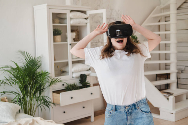 Young woman in jeans and white T-shirt wearing virtual reality helmet plays game, works, watches video in room next to bed. Concept of modern technology, VR, augmented virtual games, entertainment. - Foto, imagen