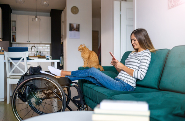 Mujer joven discapacitada usando un teléfono inteligente mientras está sentada en el sofá en casa - Foto, imagen