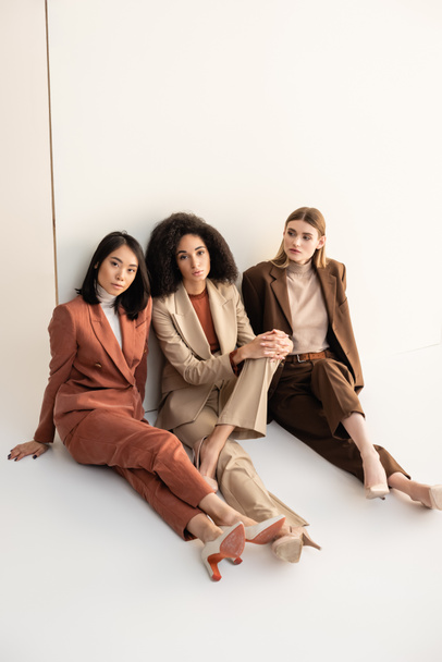 young multicultural women in trendy suits sitting and posing on white - Valokuva, kuva