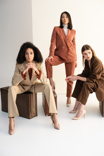full length of multicultural women in stylish suits sitting on suitcases on white - Foto, imagen