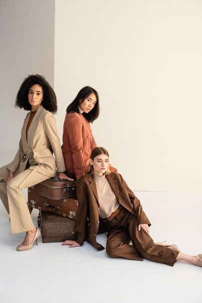 volledige lengte van jonge multiculturele vrouwen in trendy pakken zittend op koffers op wit - Foto, afbeelding