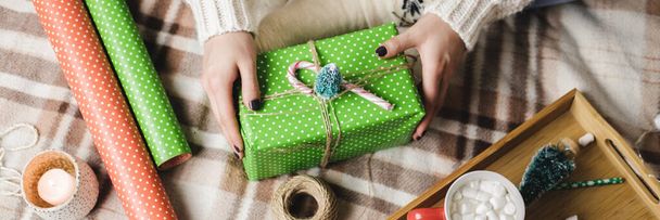 Young woman sits on plaid in cozy knitted woolen white sweater, socks and wraps Christmas gift in polka dot wrapping paper. Wooden tray with mug of cocoa with marshmallows, toy tree, candle, straws. - Fotó, kép