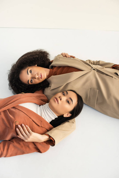 top view of asian and african american models in trendy suits lying on white  - Φωτογραφία, εικόνα