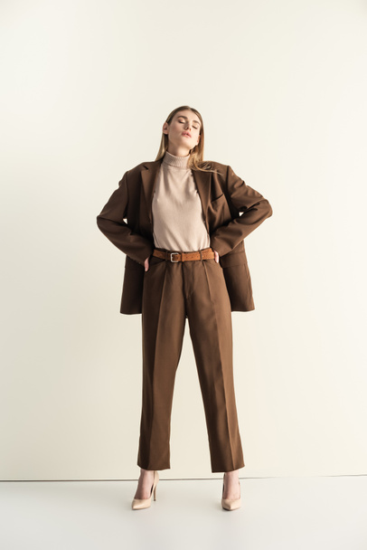 full length of young blonde woman in stylish brown suit standing with hands on hips on white - Foto, afbeelding