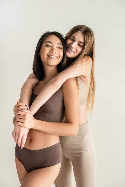 blonde woman hugging happy asian model while posing isolated on white - Zdjęcie, obraz