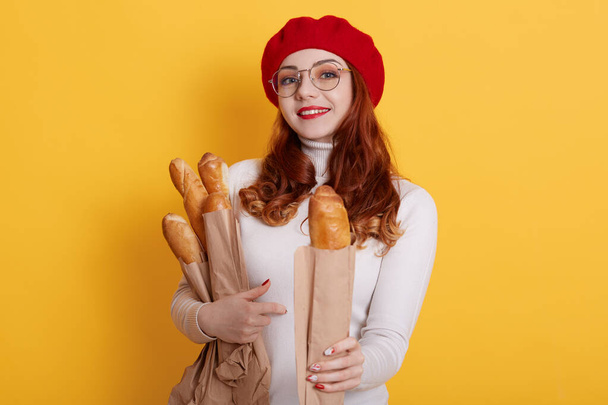 Adorable woman holding paper bag with long bread baguettes, offers one to somebody, wearing white shirt, red beret and glasses, wears red lipstick, against yellow background. - Foto, imagen