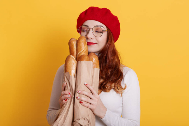 Beautiful red haired young woman holding paper bag with bread on yellow background, satisfied girl smells fresh fragrant long loaf with closed eyes, lady wearing casual shirt and beret. - Фото, зображення