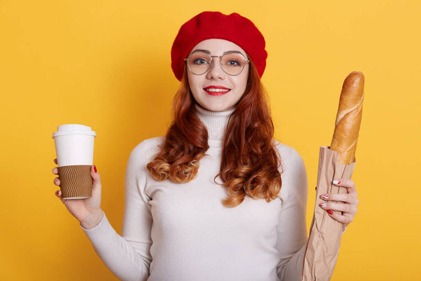 Portrait of girl with long red hair wearing beret, white shirt and glasses, looks at camera and smiles, holding bread in paper bag and take away coffee, model posing against yellow wall. - Foto, afbeelding