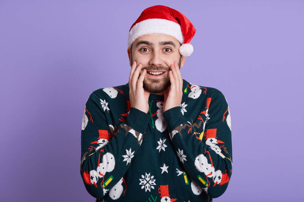 Bearded guy dresses santa hat and fanny sweater with snowman looking at camera, stands with hands on cheeks, being surprised by shocking news, posing isolated over lilac background. - Foto, Imagen