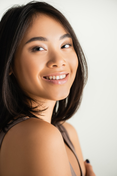 brunette asian woman smiling while looking at camera isolated on white - Zdjęcie, obraz