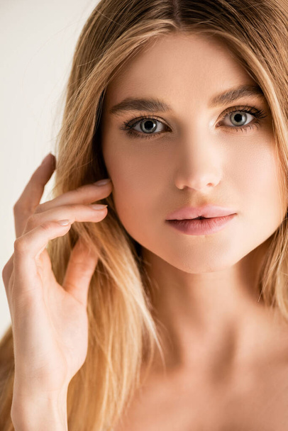 young blonde model looking at camera while fixing hair isolated on white - Φωτογραφία, εικόνα