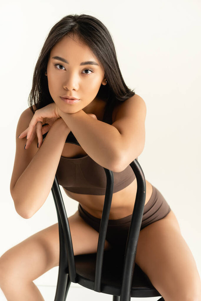 brünette asiatische Frau in beiger Unterwäsche schaut in die Kamera, während sie auf einem Stuhl isoliert auf weiß posiert - Foto, Bild