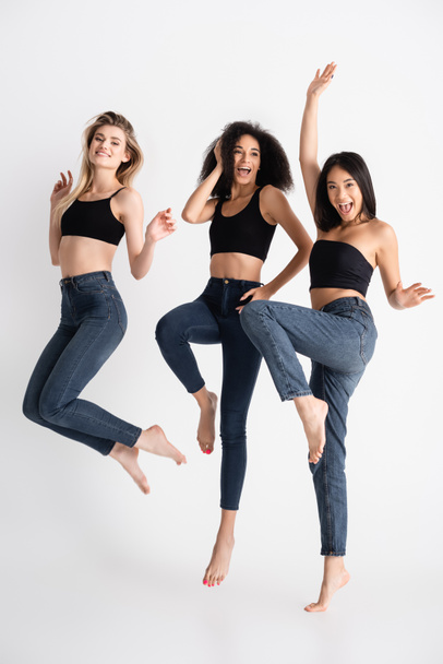 excited interracial women in denim jeans jumping and posing on white - Foto, Imagen