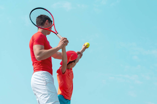 A young tennis coach or instructor teaches a child a serving technique. Boy tennis player throws the ball up for serve. Kids sports school. Blue sky background. Banner. Copy space for text. - Zdjęcie, obraz