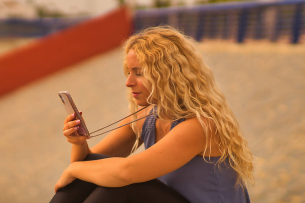 Young blonde woman consulting social networks on her smartphone sitting on a park bench. - Foto, imagen